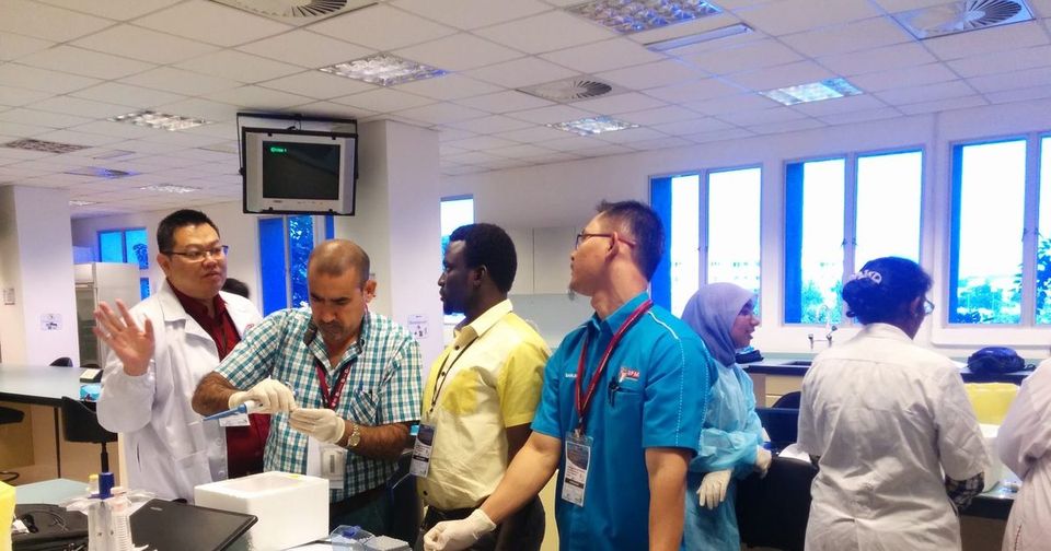 A man in a lab coat explains a step to a group standing at a laboratory bench