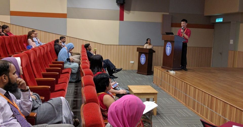 A speaker at the lectern on stage while delegates listen from the red seats of the auditorium.