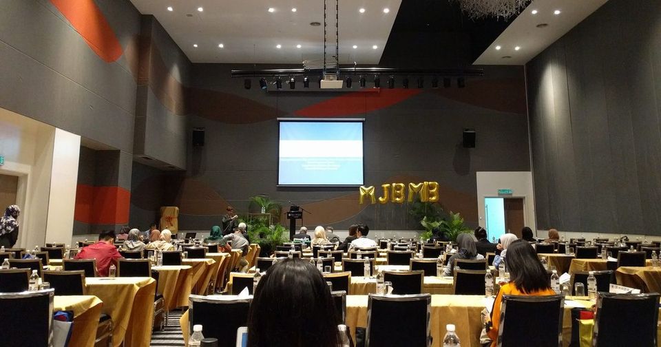 Delegates seated at long tables in the conference hall, facing a screen and a stage with MSBMB balloon letters
