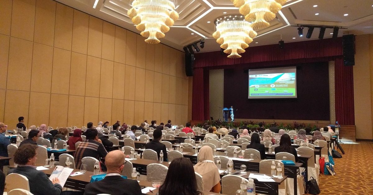 Delegates seated at long tables in a large hotel ballroom, facing the stage and a screen