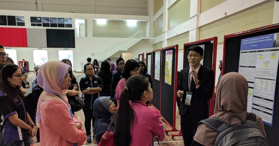 A presenter in a dark suit explains his research poster to a group of visitors in the hall