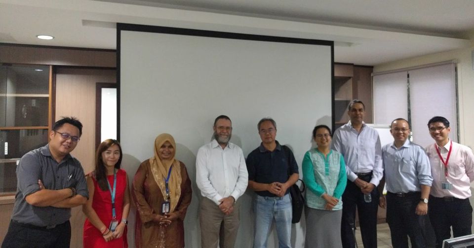 Members and guests stand in a row for a group photo in a meeting room, in front of a screen
