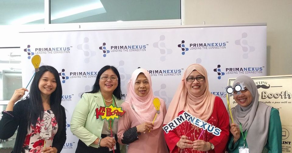 Five women hold photo props in front of the Prima Nexus backdrop at the photo booth