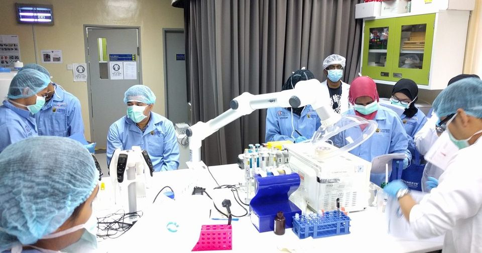 Participants in lab coats, masks and hair covers gather around a laboratory bench with pipettes and sample racks