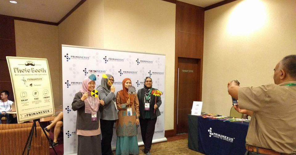 Four delegates hold photo props in front of the Prima Nexus backdrop while a man takes their picture
