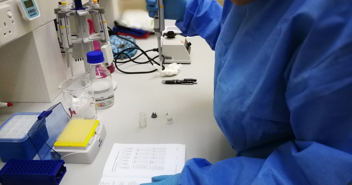 Gloved hands hold a pipette over a kit protocol at a lab bench, beside tube racks, reagent vials and a well plate.