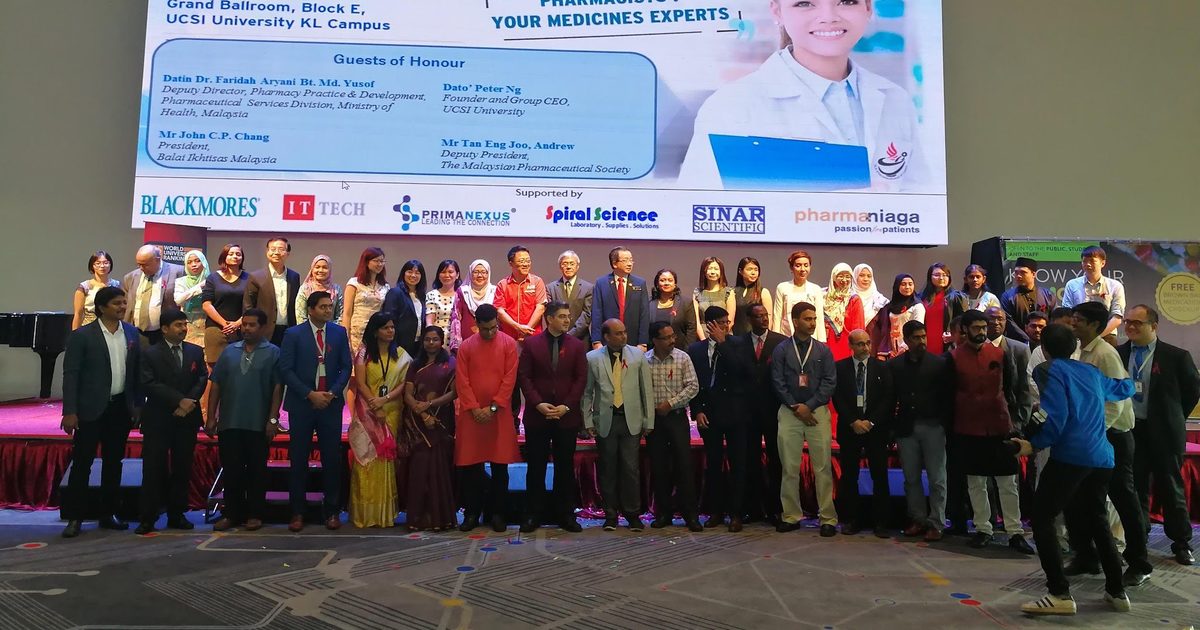 Students, staff and guests in a group photo on stage below the World Pharmacists Day screen