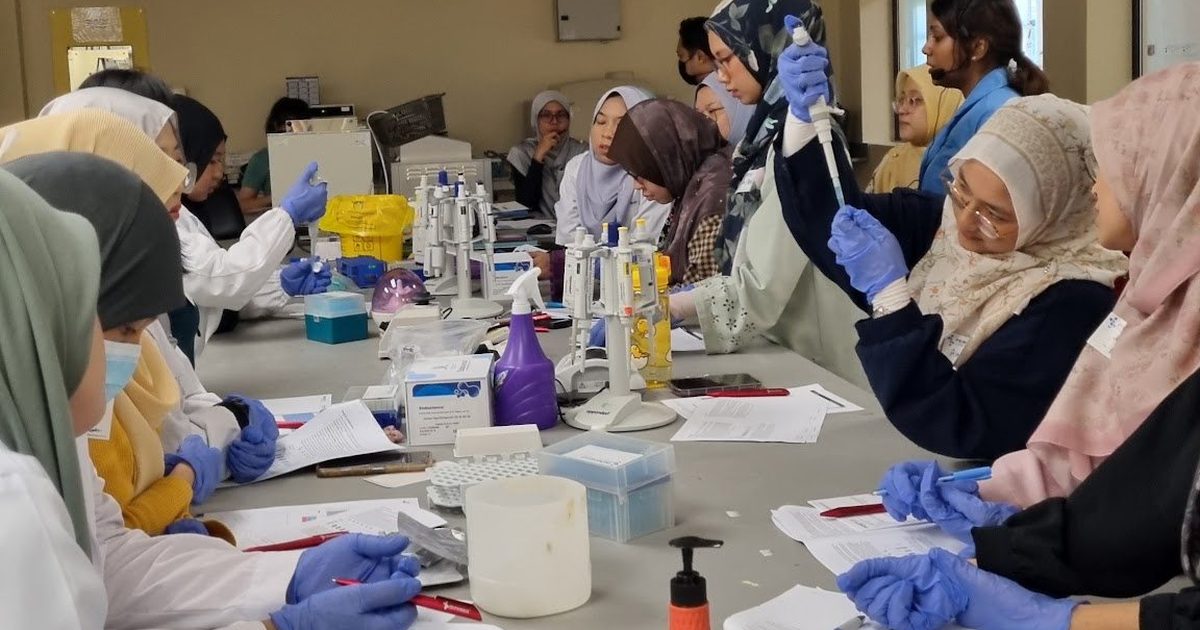 Participants in lab coats and gloves at long laboratory benches, one raising a hand
