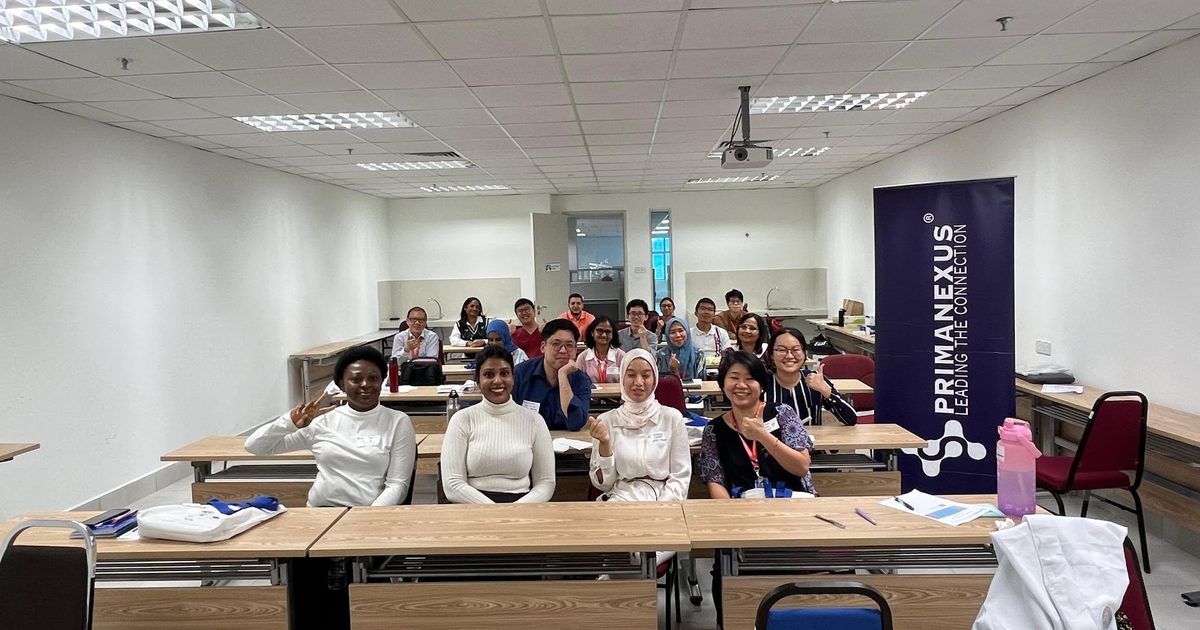 Workshop participants seated at tables in a classroom for a group photo, beside a Prima Nexus banner