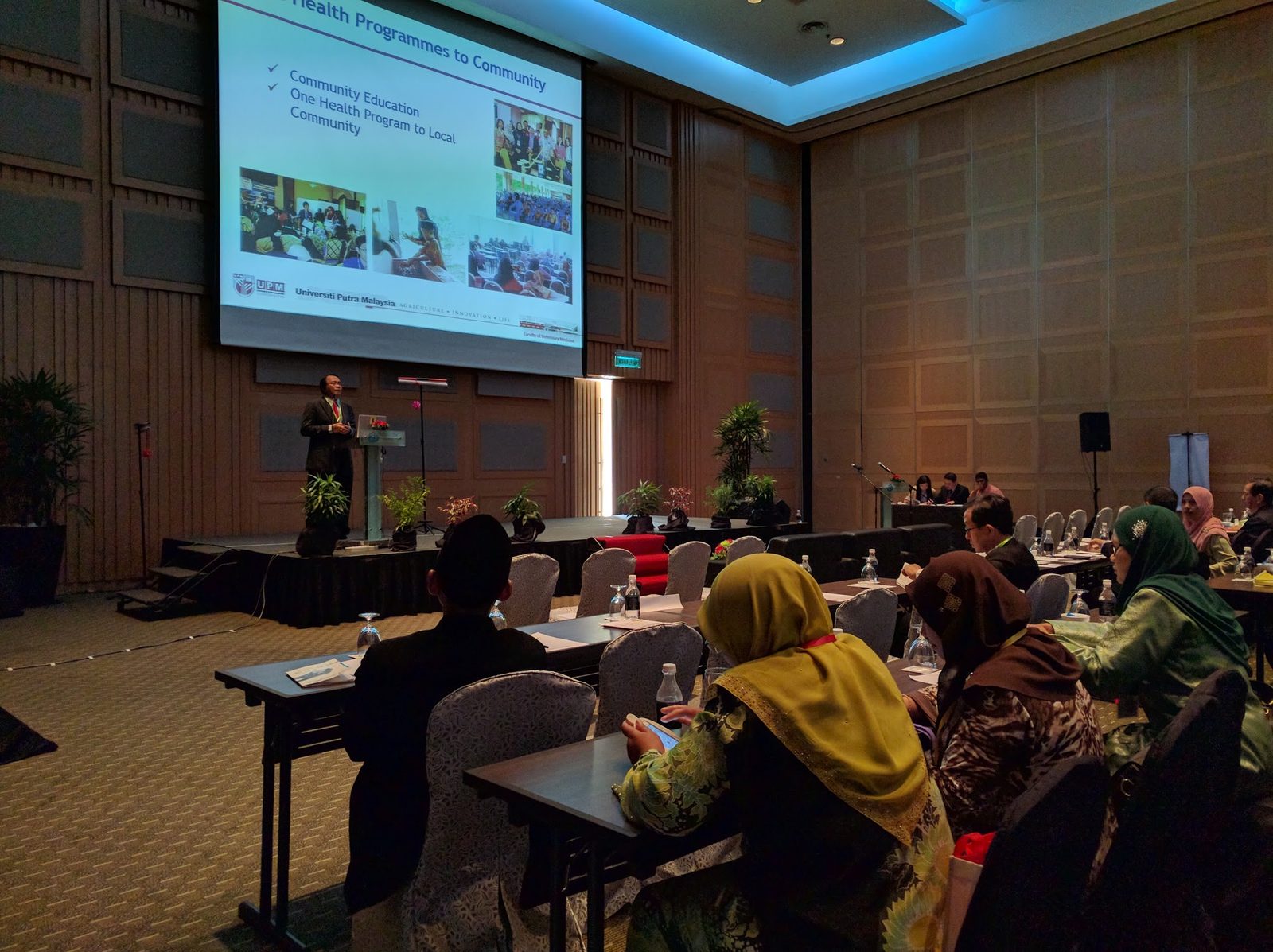A speaker presents to delegates seated at long tables in a conference hall, with slides on a large screen