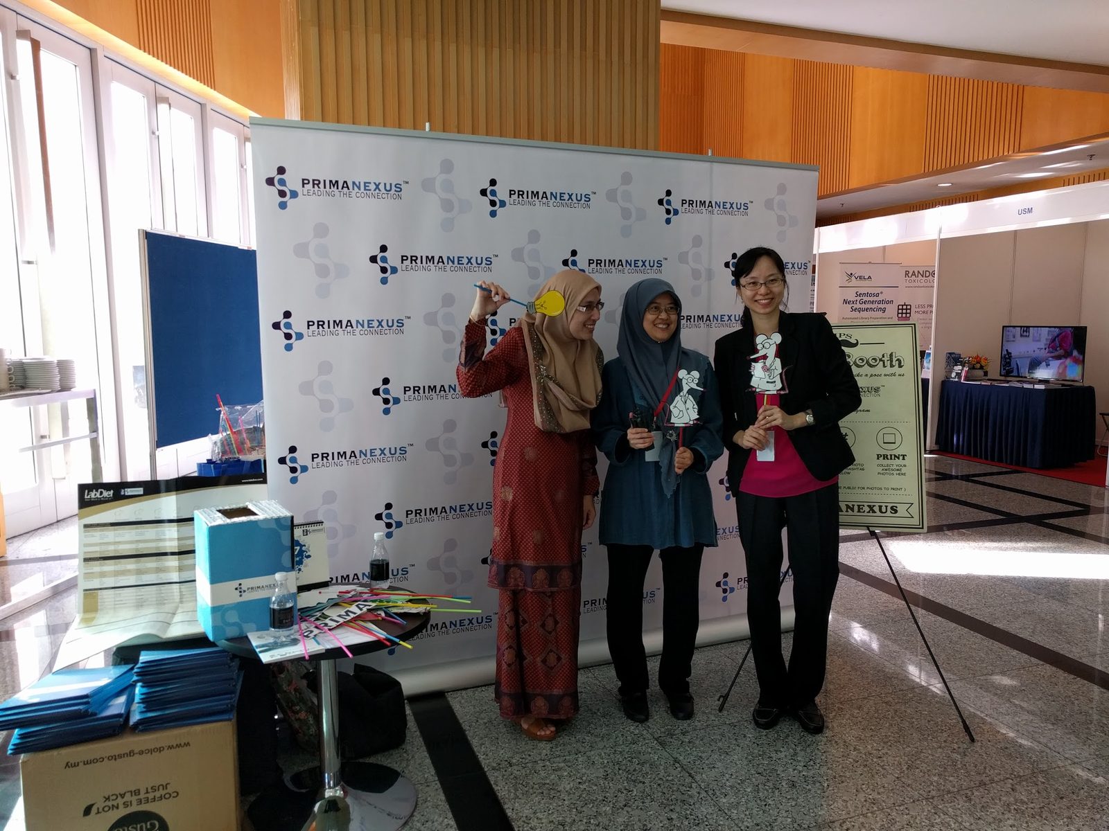 Three delegates with photo props at the Prima Nexus photo booth, with blue folders stacked on a table beside it
