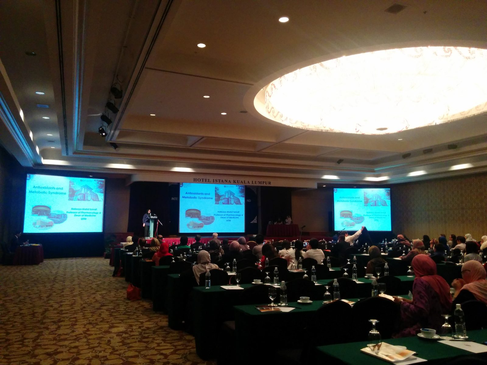 A speaker at the lectern, with her talk title on antioxidants and metabolic syndrome shown on three large screens
