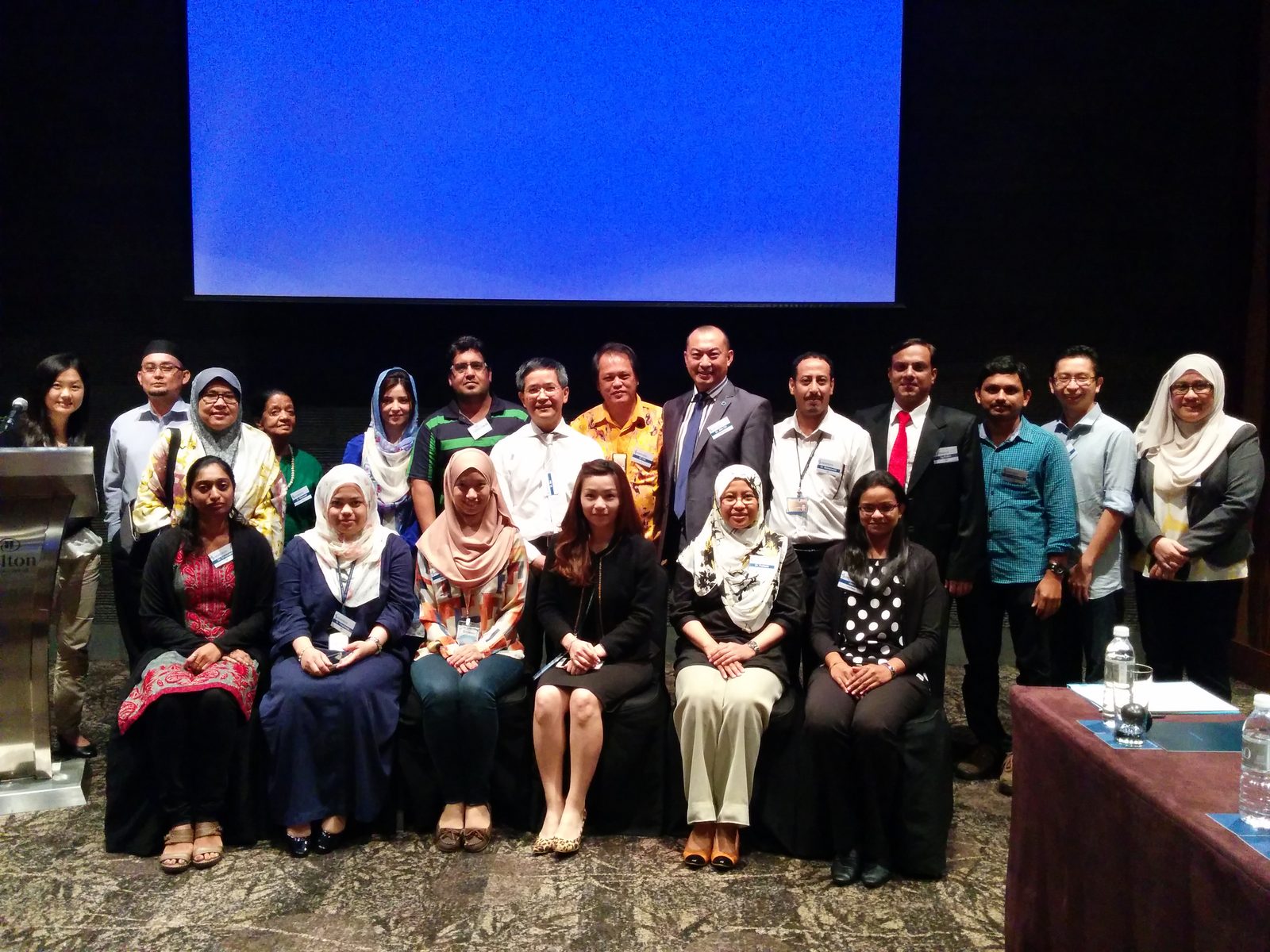 Group photo of participants standing and seated in front of the screen at Hilton Hotel, KL Sentral