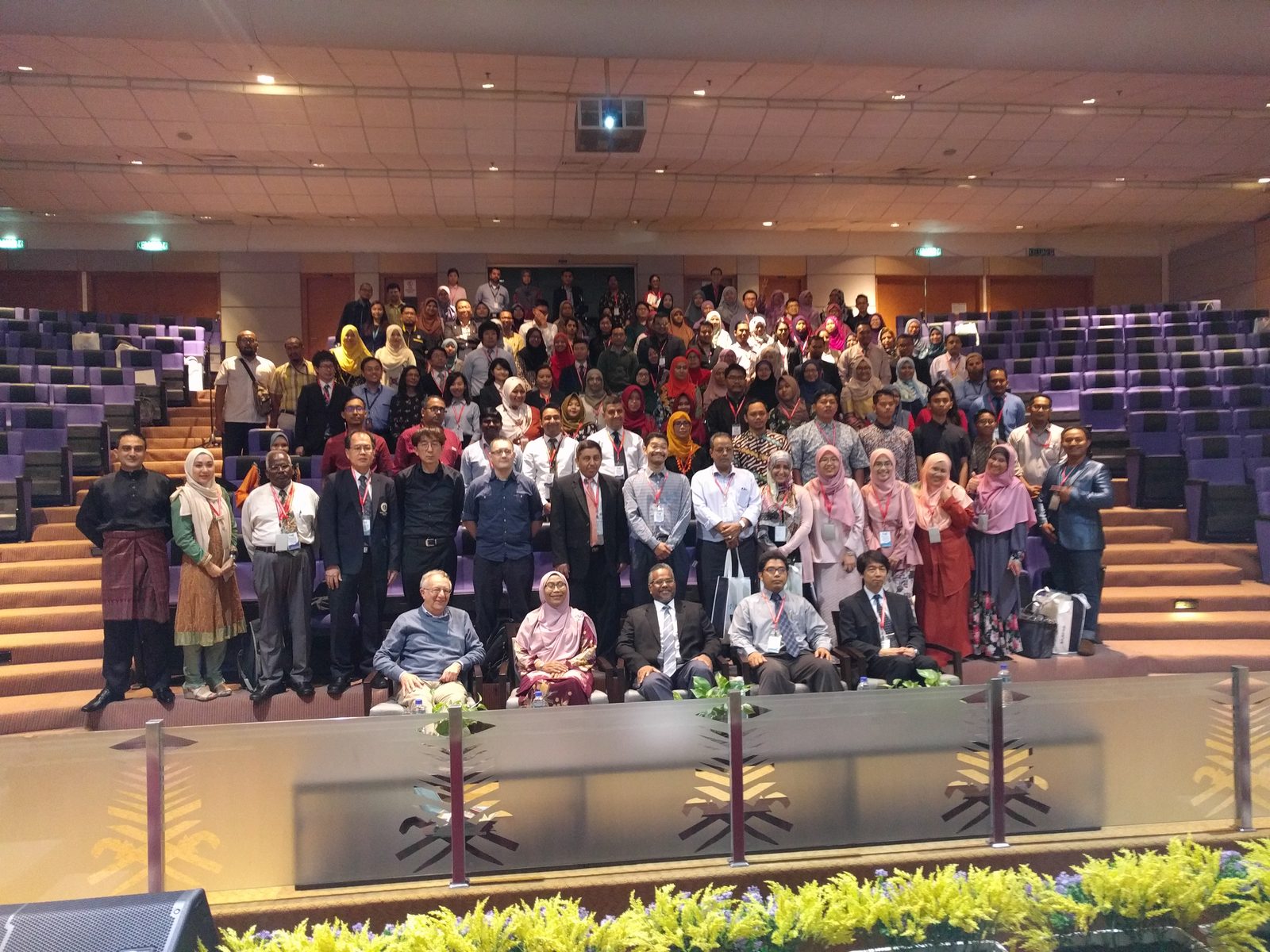 Large group photo of conference participants on the tiered seats of a lecture hall, front row seated
