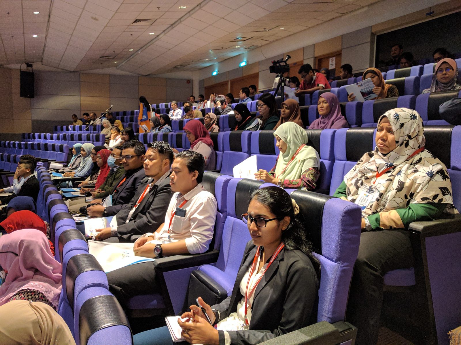 Delegates seated in rows of purple seats in a lecture hall, following a conference session