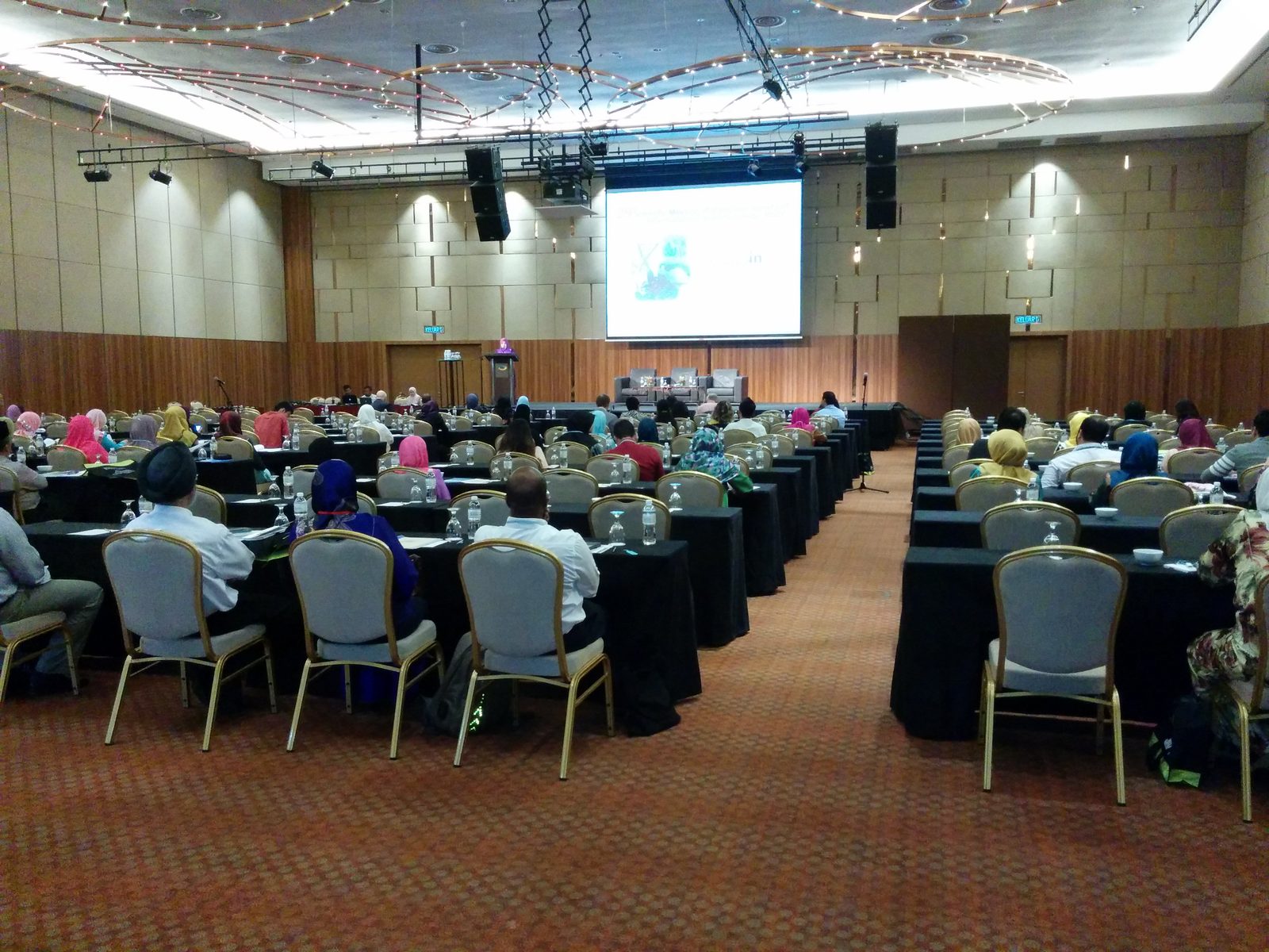 Delegates seated at rows of long tables in the ballroom, facing the stage and a large screen