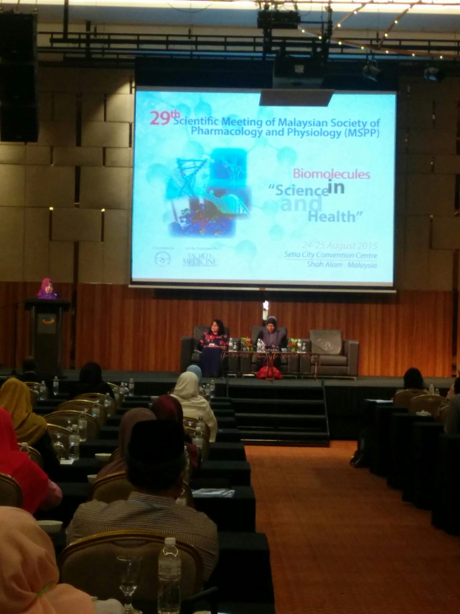 A session on stage, with the meeting's title, theme, dates and venue shown on the screen above