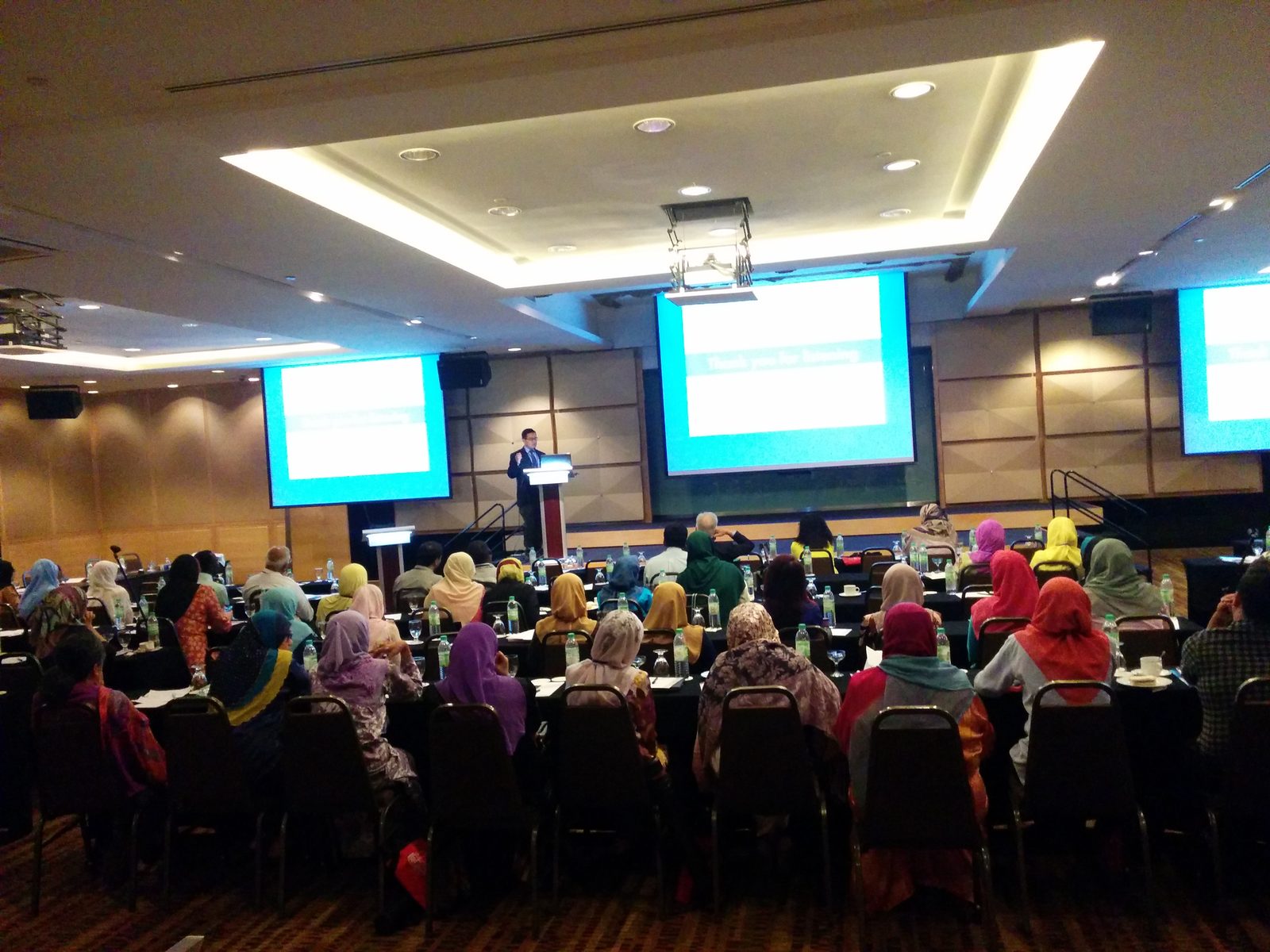 Delegates seated at long tables in a hotel ballroom, facing a speaker at a lectern and three large screens