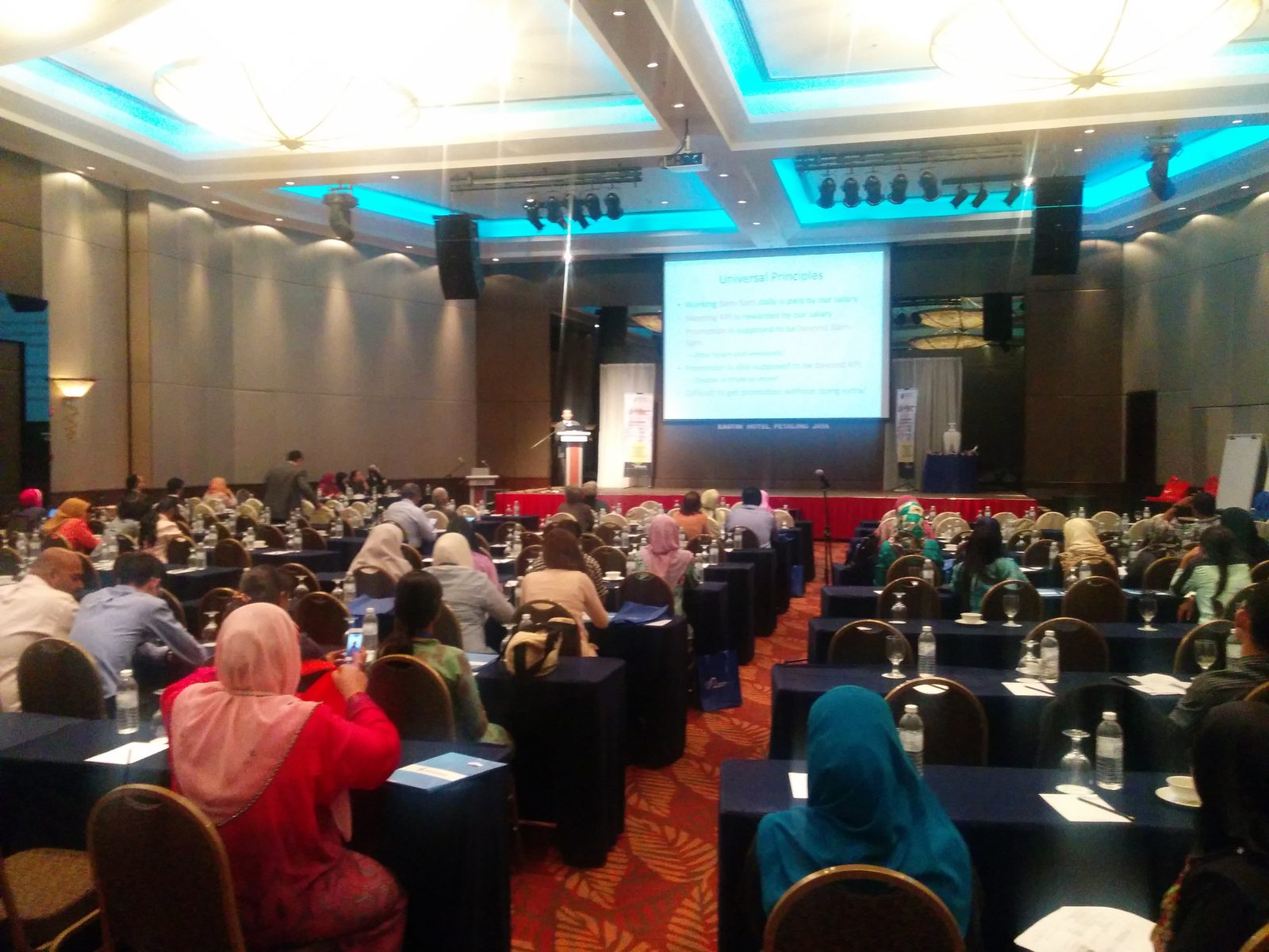 Delegates seated at long tables in a hotel ballroom, facing the stage and a projected lecture slide