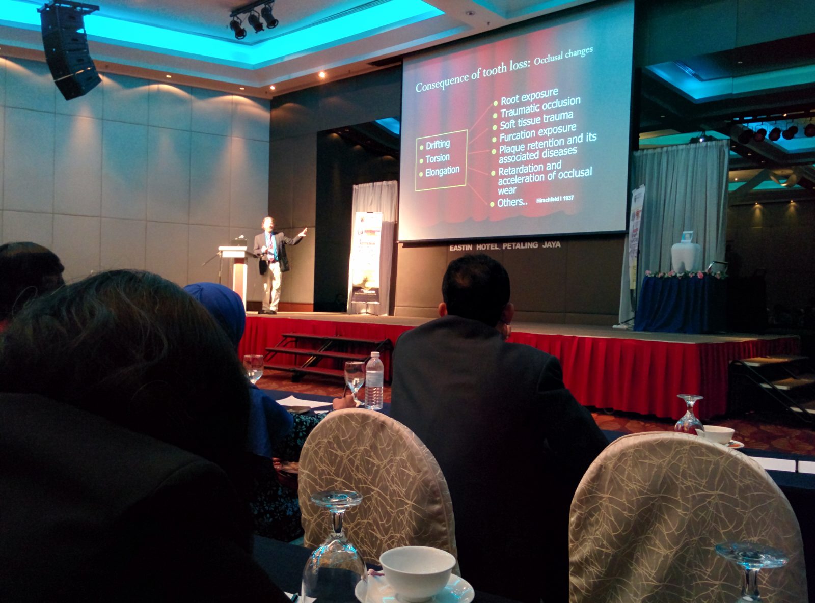A speaker on stage presents a slide on the consequences of tooth loss to a seated audience