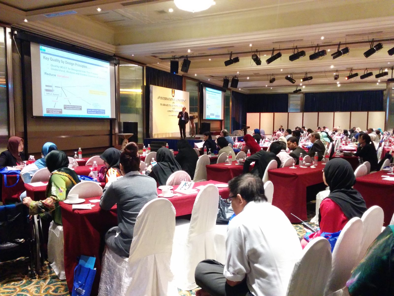 Delegates seated at round tables in the conference hall while a speaker presents from the front