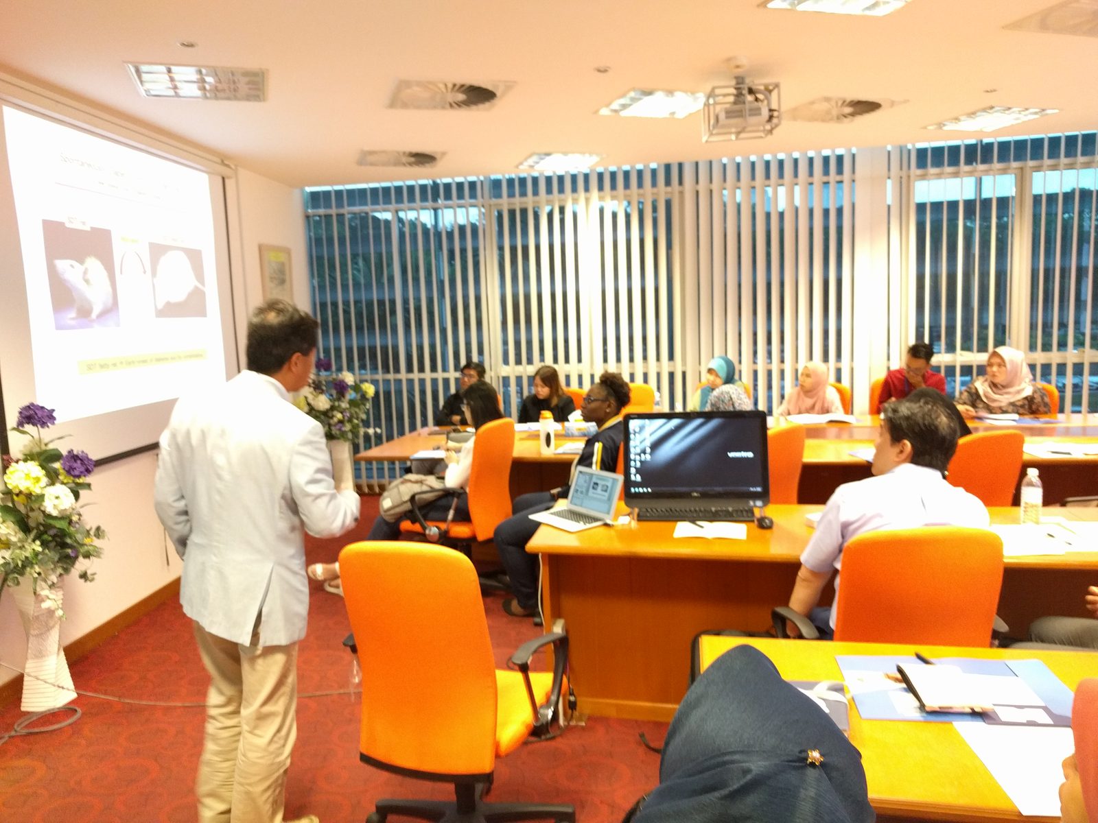 A speaker in a light jacket presents a slide showing laboratory rats to participants seated at desks.
