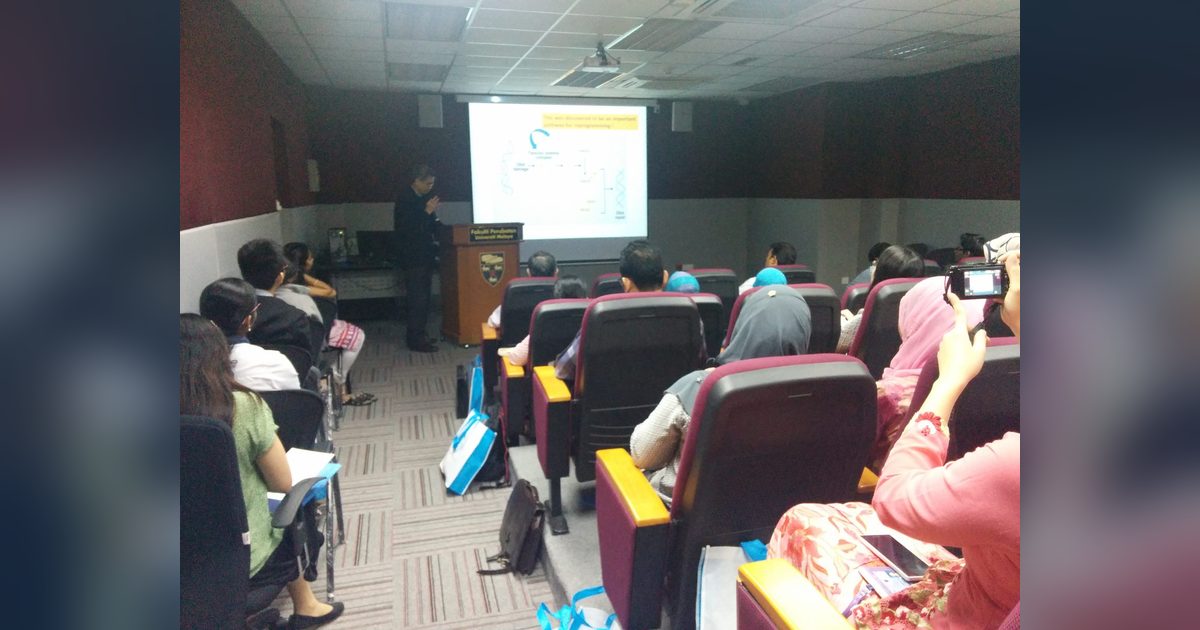 A speaker at a lectern presents a slide to a seated audience in a lecture hall