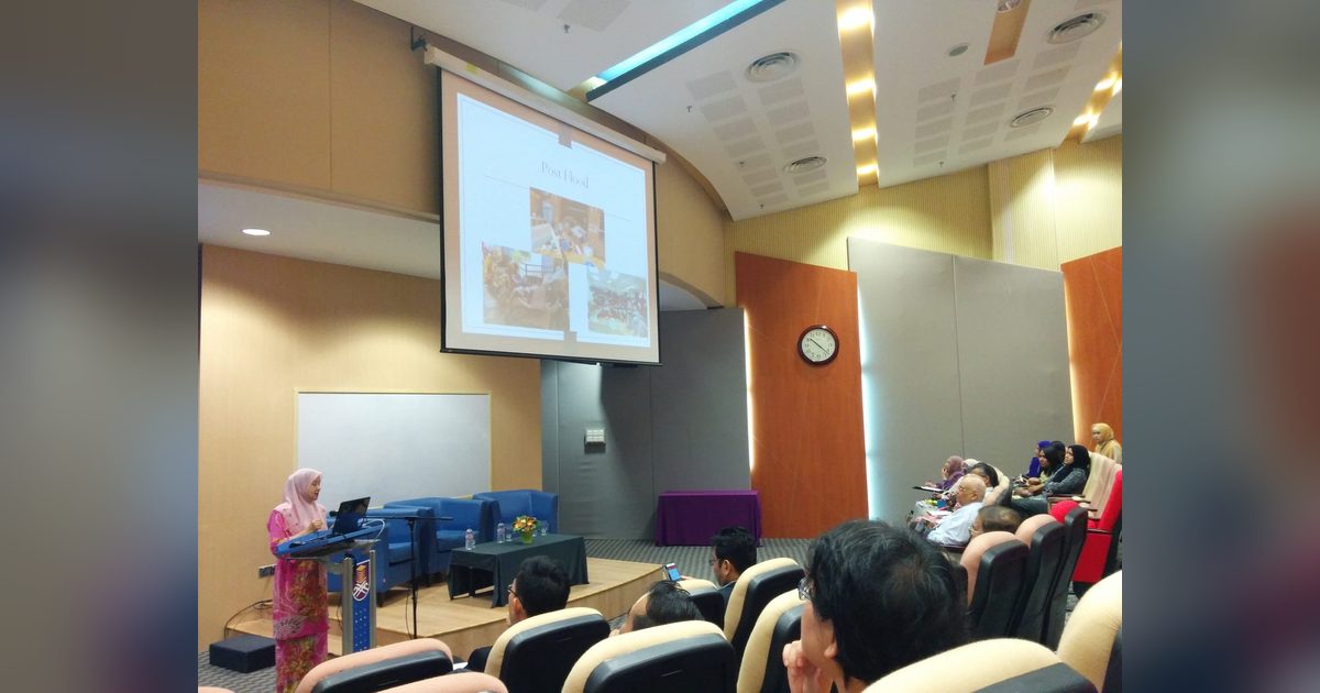 A speaker at the lectern presents a slide titled Post Flood to an audience in a university auditorium
