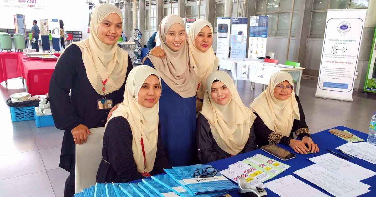 Six women at a registration desk with a stack of blue Prima Nexus folders on the table in front of them