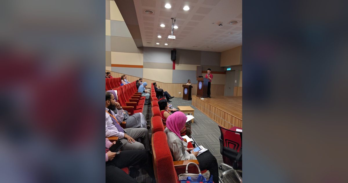 A speaker at the lectern on stage while delegates listen from the red seats of the auditorium.