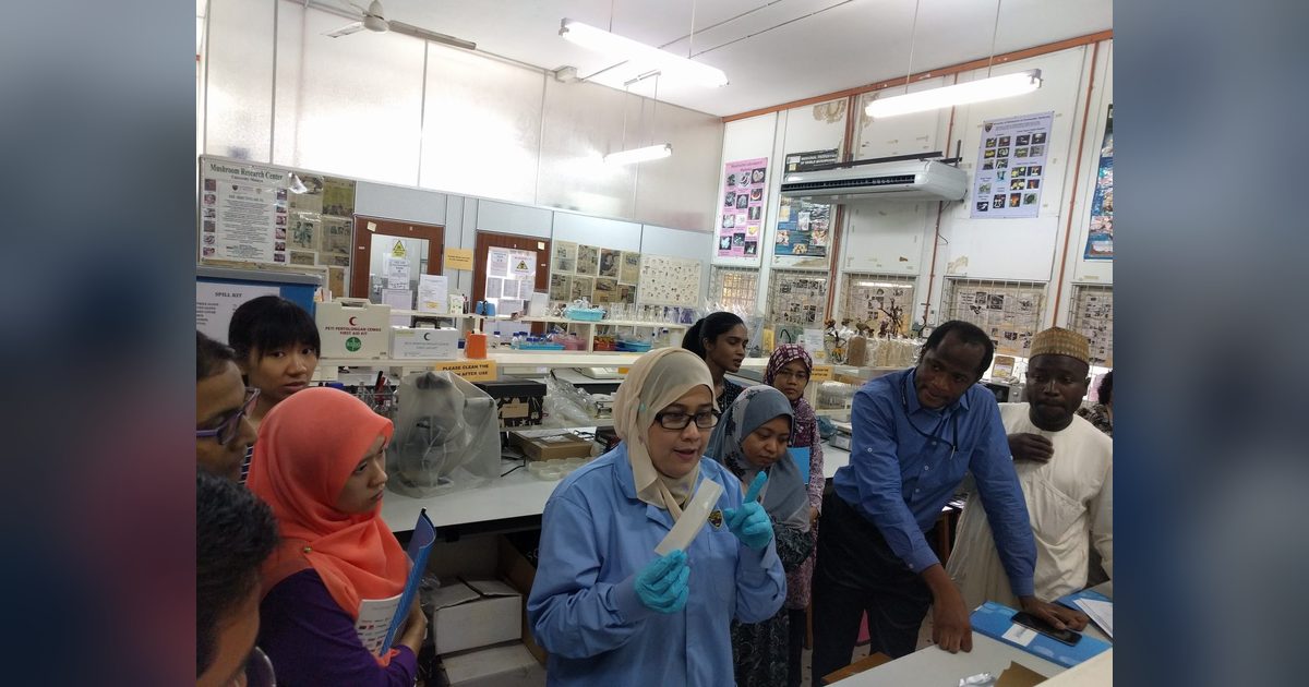 A demonstrator in a blue lab coat and gloves holds up an assay item and explains it to participants at a lab bench.