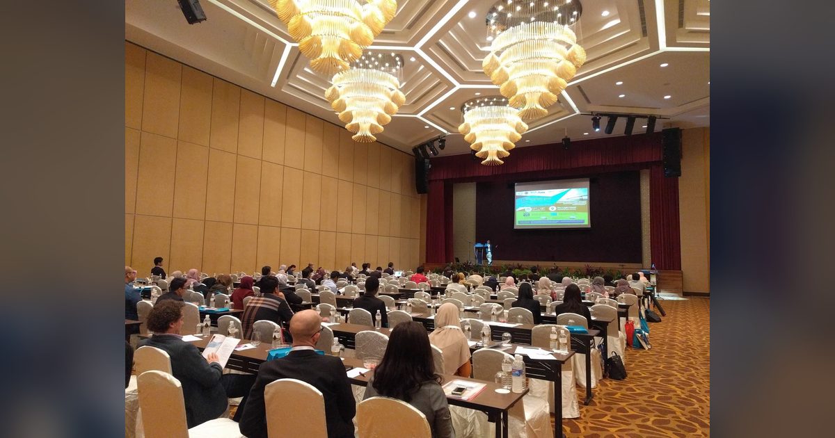Delegates seated at long tables in a large hotel ballroom, facing the stage and a screen