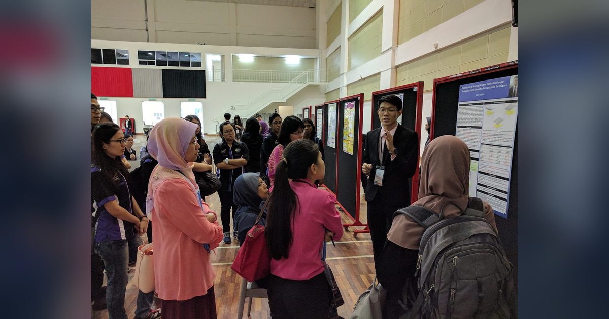 A presenter in a dark suit explains his research poster to a group of visitors in the hall