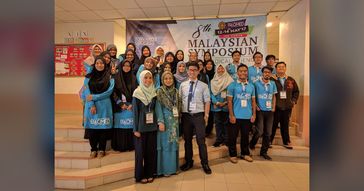 A large group, many in blue symposium T-shirts, poses in front of the 8th Malaysian Symposium banner.