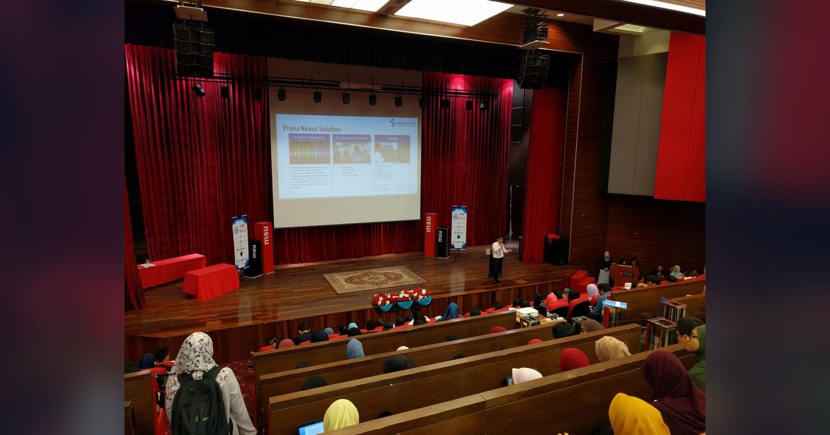 Speaker on the theatre stage with a Prima Nexus slide on the screen and students seated in rows