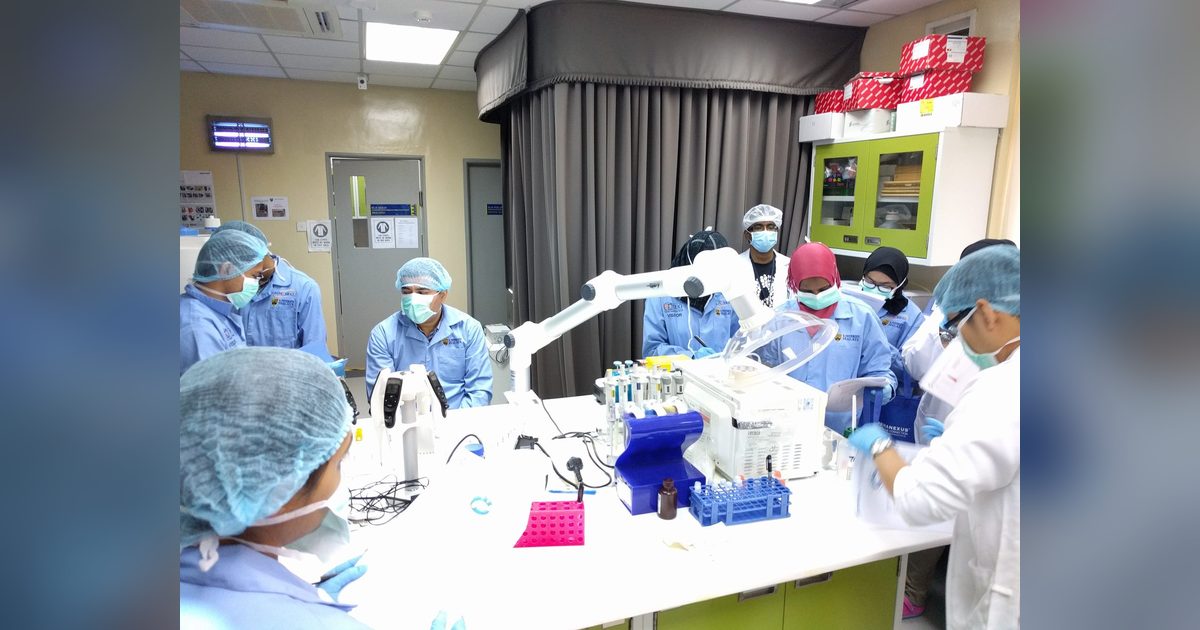 Participants in lab coats, masks and hair covers gather around a laboratory bench with pipettes and sample racks