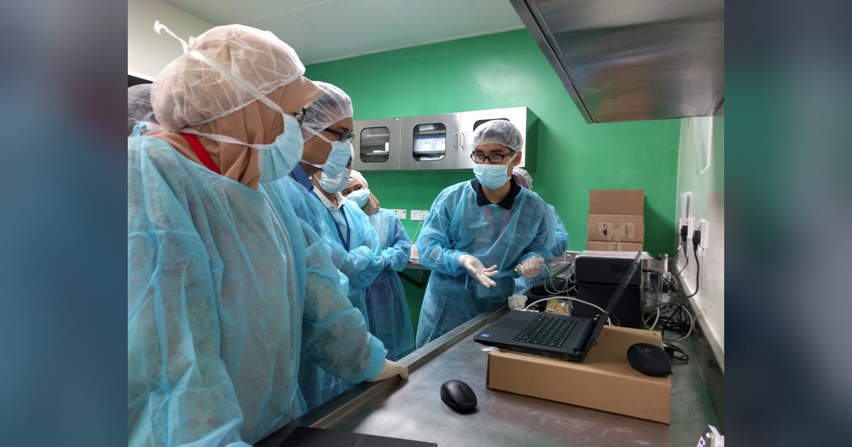 A trainer in gown and mask demonstrates the CODA system at a lab bench to a group of gowned participants