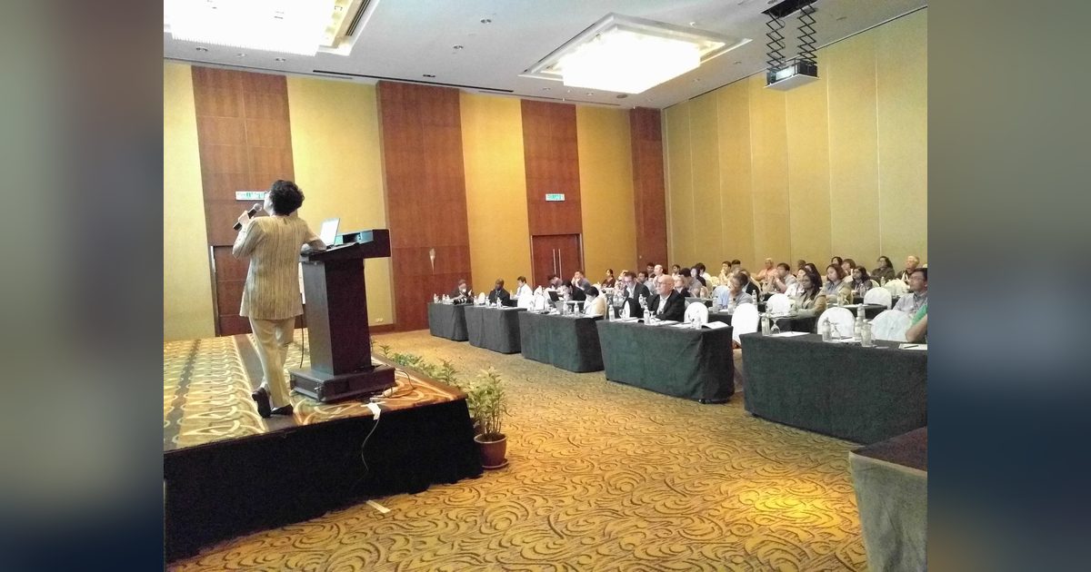 A speaker at the lectern addresses delegates seated at long tables in the conference hall