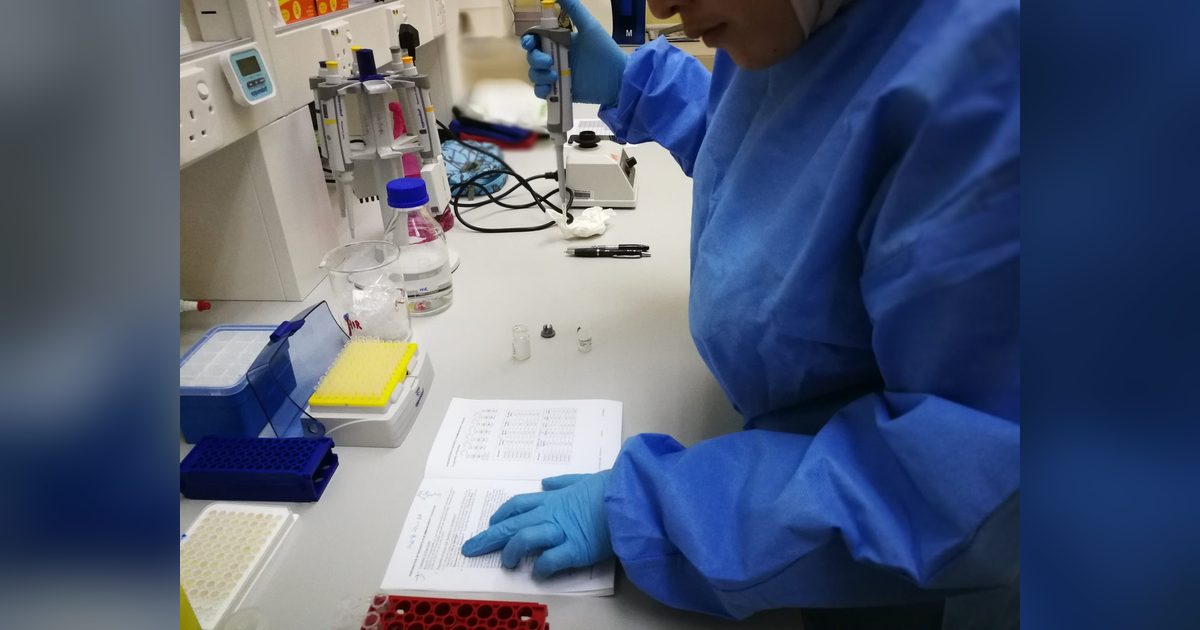 Gloved hands hold a pipette over a kit protocol at a lab bench, beside tube racks, reagent vials and a well plate.