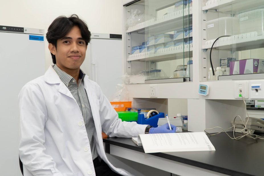 A scientist in a lab coat seated at a laboratory bench with a printed document, freezers and shelves of supplies behind him