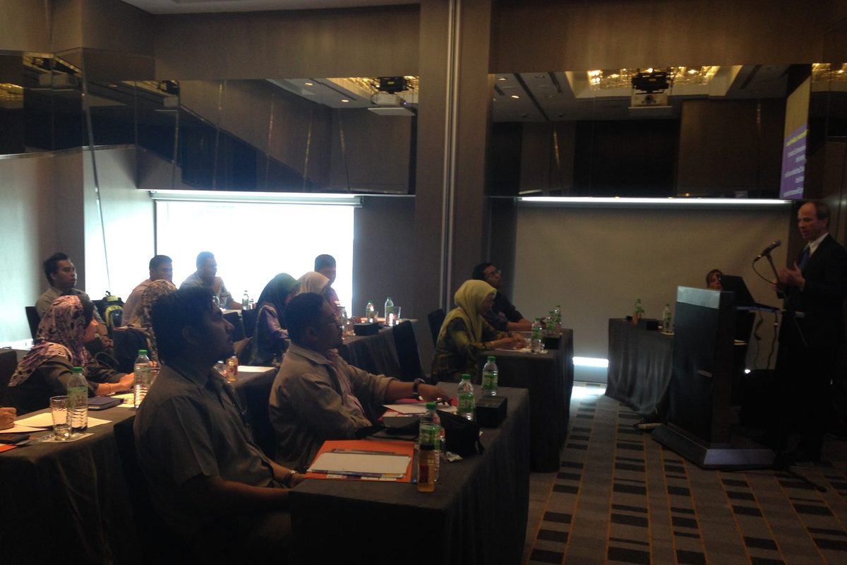 Participants seated at tables in a hotel meeting room, facing a speaker at a lectern