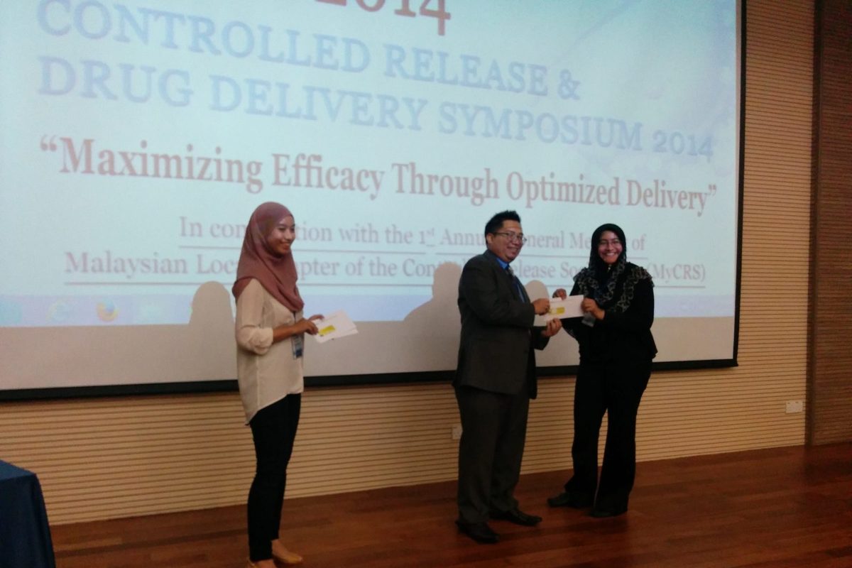 A man hands an envelope to a woman on the stage while another woman stands beside them, in front of the symposium slide