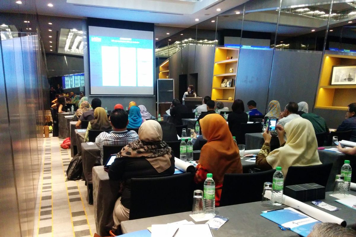 Participants seated at long tables in a hotel meeting room, watching a presentation on a large screen