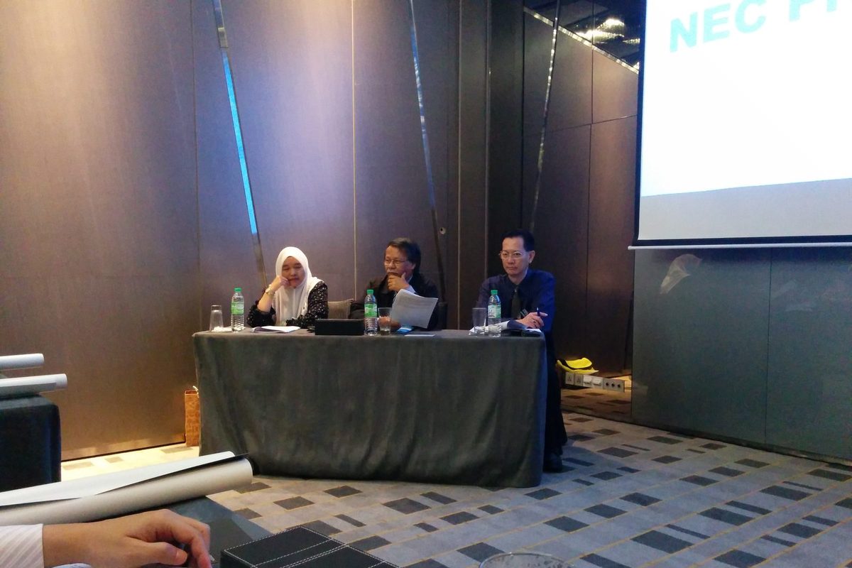 Three people seated at a head table at the front of a meeting room, with a projection screen to one side