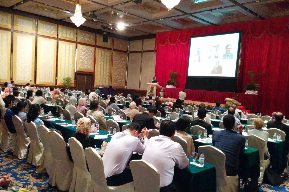 Delegates seated at long tables in a large hall with red curtains, facing a speaker and a screen