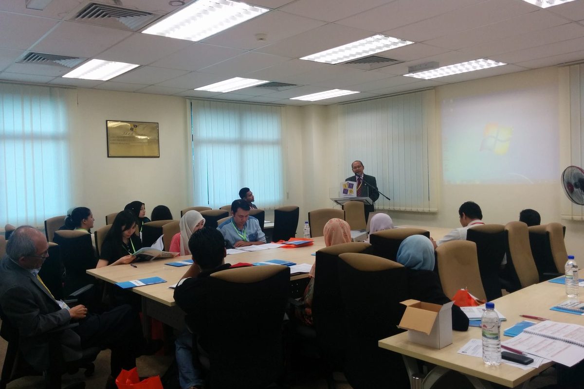 Participants seated at long tables in a seminar room, facing a speaker at a lectern