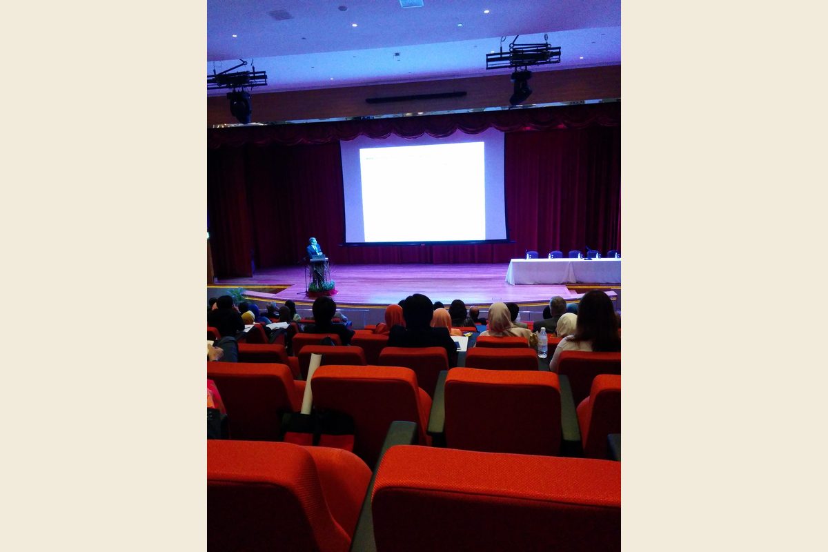Delegates in red auditorium seats facing a speaker at a lectern and a large screen