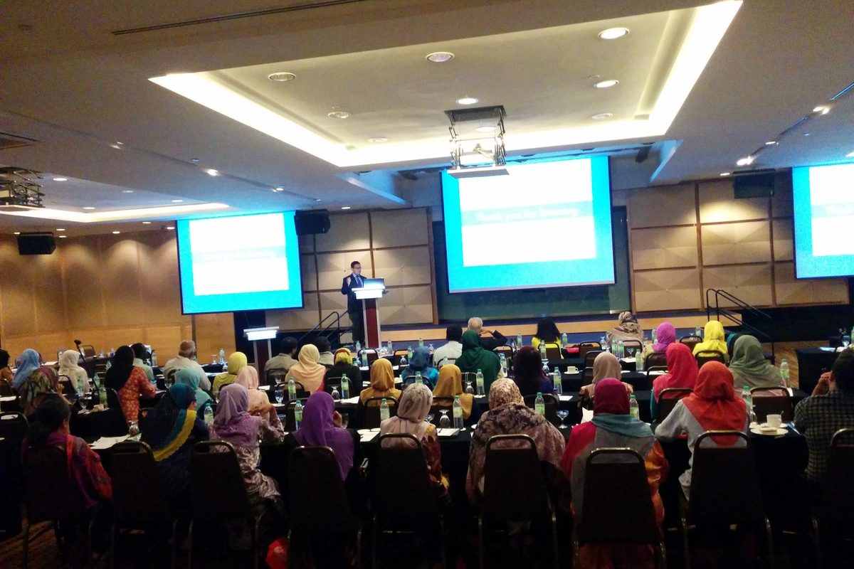 Delegates seated at long tables in a hotel ballroom, facing a speaker at a lectern and three large screens