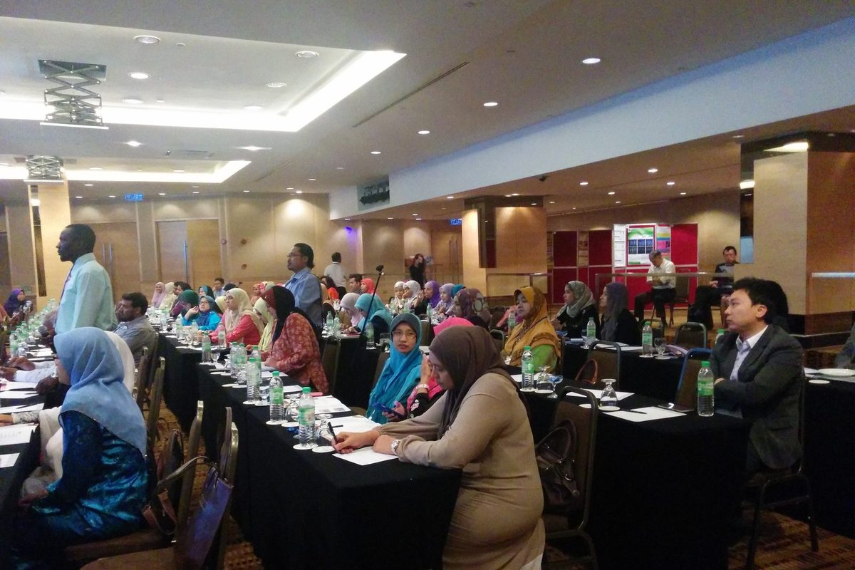 Delegates seated at long tables in a hotel function room, with poster boards at the back
