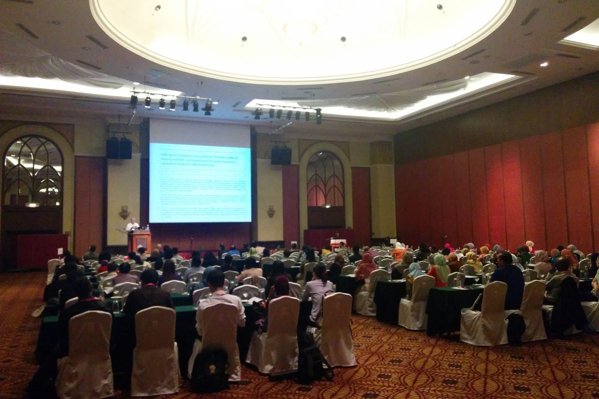 Delegates seated at round tables in a hotel ballroom, facing a stage and a large screen